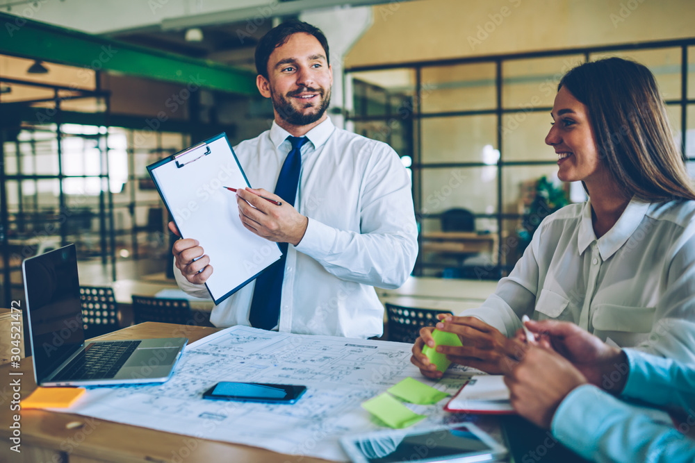 Poster smiling businessman showing paper folder to colleagues sitting near desktop with blueprint and lapto