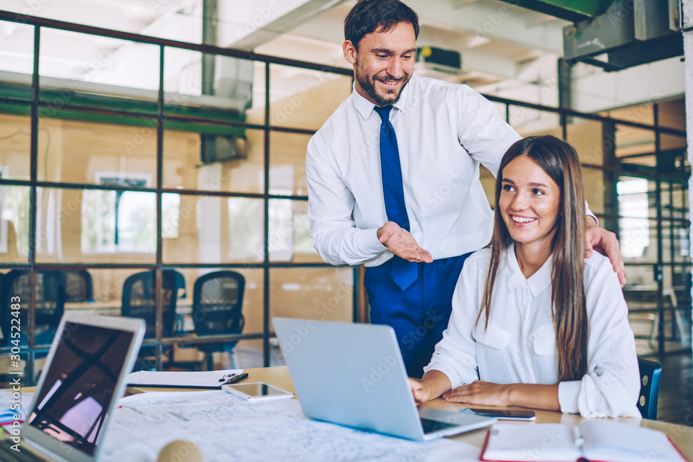 Poster smiling businessman talking to female colleague during break near desktop with laptop computer, succ