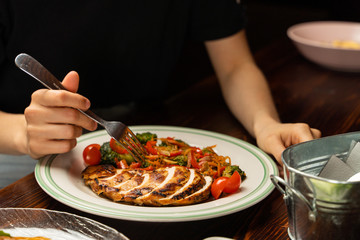 Young woman is eating grilled chicken breast with stewed vegetables:broccoli, carrot, tomatoes and bell pepper, alone with hands visible