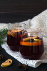 winter still life with tasty mulled wine in a two glasses with orange and cinnamon, anise star and warm white plaid on a dark wooden background with green pine branch. close up