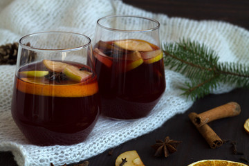winter collection. tasty mulled wine in a two glasses with orange and cinnamon, anise star and warm white plaid on a dark wooden background with green pine branch