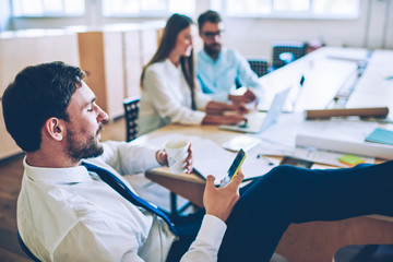 Fototapeta premium Male employee enjoying coffee break for chatting with friends via smartphone gadget while colleagues on blurred background working on laptops, businessman using cellular phone for browsing internet