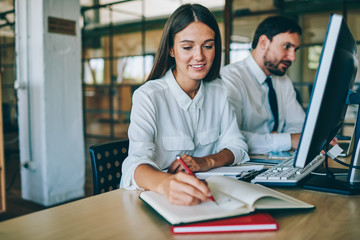 Positive woman dressed in formal wear writing information from internet in textbook sitting at desktop with modern computer, successful female employee making notes of ideas for startup project
