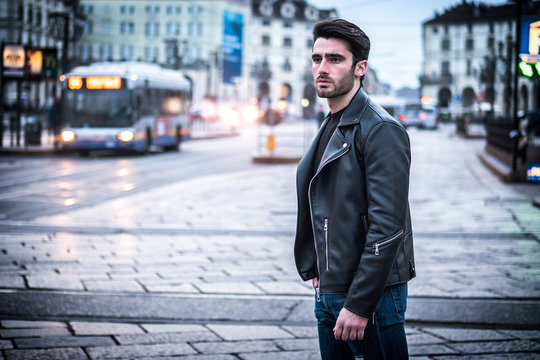 One handsome young man in urban setting in modern city, standing, wearing black leather jacket and jeans, looking at camera