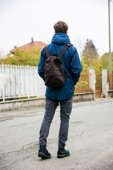 Teenage boy walking alone in city street with backpack, seen from the back