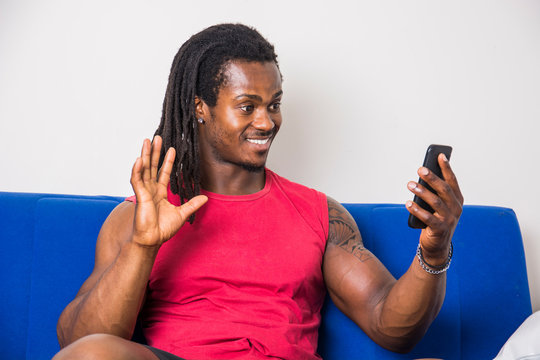 Handsome muscular black man sitting on couch at home doing videocall or videochat on cell phone, looking confident and waving with cute smiling expression