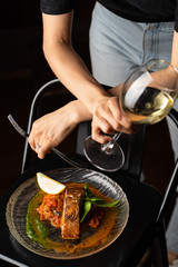 Woman is standing at the table with marinated chum salmon on it,  holding a glass of white wine and fork