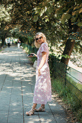 Beautiful stylish blonde woman in polka dotted midi dress in city summer park. She is happy and enjoying walk in sunny day. She is wearing small white bag and black sunglasses.  