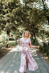 Obraz na płótnie Canvas Beautiful stylish blonde woman in polka dotted midi dress in city summer park. She is happy and enjoying walk in sunny day. She is wearing small white bag and black sunglasses. 