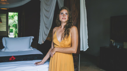 Adult woman pondering while standing near bed in cozy bungalow