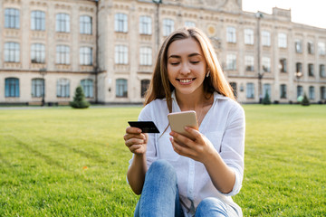 Pretty young woman student sitting in street city park and doing online shopping, using online...