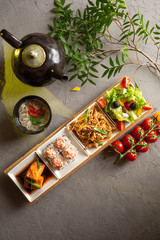 Complete lunch. Greek salad, glass noodles, rolls, salmon fillet, noodle soup and teapot with tea on a gray background.