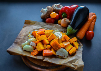 Autumn vegan food: baked pumpkin with carrots, onions and spices on baking paper. In the background vegetables: mushrooms, carrots, red bell pepper, tomatoes, onion and eggplant. On black background