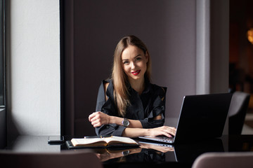 Positive young business woman sitting on workplace with laptop and diary.