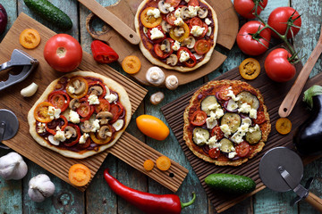 Homemade vegetarian pizza party. Flat lay of gluten free cauliflower and dough pizzas with eggplants, mushrooms, tomatoes and goat cheese decorated with raw produce on oak boards