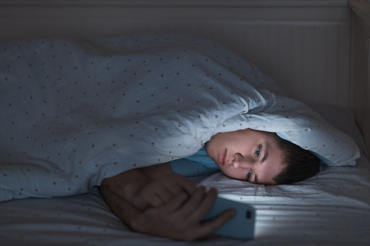 Sleepy depressed teenager surfing in the internet on his mobile phone lying on bed in the dark. Web addiction, smartphone dependency, internet trap. Insomnia, nomophobia, sleep disorder concept.