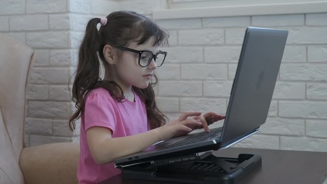 The child is at the computer. Cute little girl in glasses is typing on a laptop.ce.
