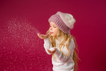 little blonde girl in a knitted hat and sweater catches the snow and laughs. winter clothes. isolate on red background, space for text