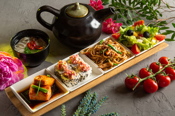 Complete lunch. Greek salad, glass noodles, rolls, salmon fillet, noodle soup and teapot with tea on a gray background.