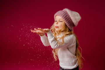 little blonde girl in a knitted hat and sweater catches the snow and laughs. winter clothes. isolate on red background, space for text
