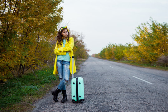 girl with a suitcase is walking along a road. The girl with wavy hair returns home. Young woman with a suitcase. Beautiful girl in a yellow raincoat in the fall. Hitchhiking on the road with luggage
