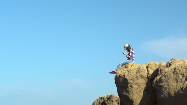 A young woman is sitting on a rock with a telephone. A woman in a cap dangled her legs over a cliff. Communication in the messenger on a smartphone