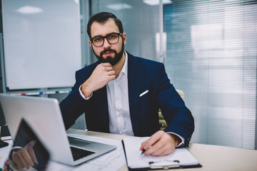 Half length portrait of successful proud ceo dressed in formal wear looking at camera while writing down financial reports and working at modern laptop computer sitting in office of corporate company