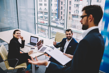 Positive male and female entrepreneurs in formal wear sitting at meeting table with laptop computers and attentively listening leader of financial company during working briefing in office