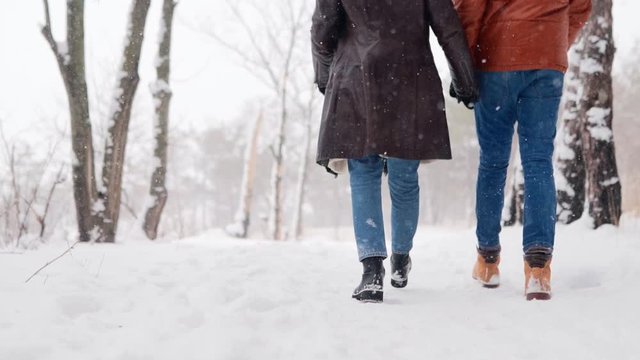 Back view of loving couple feet walking in a park on snowfall. Man and his girlfriend holding hands enjoying snow on winter day. Hipster woman stroll in snowy forest with boyfriend in stylish clothing