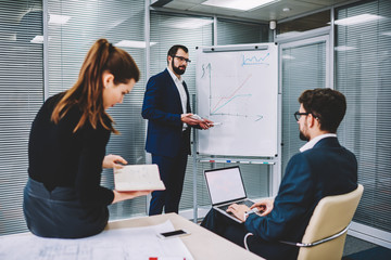 Experienced executive manager in spectacles standing next to flip chart and explaining for colleagues holding laptop computer with blank copy space screen for your advertising information content