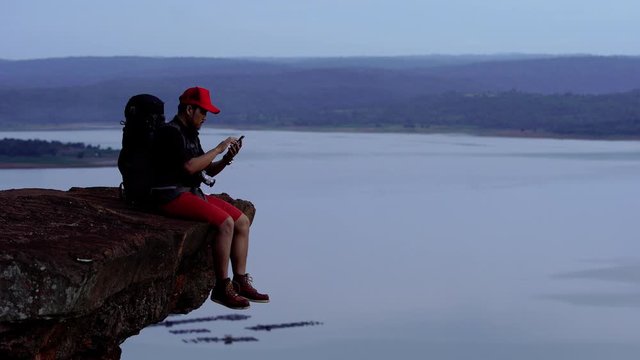 hiker man sitting and using smartphone on the edge of cliff, on a top of the rock mountain