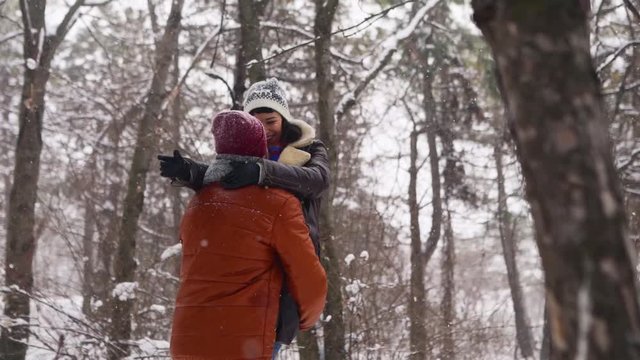 Man spinning or turning happy girlfriend holding her in his hands on snowfall in pine forest. Loving couple have fun in snowy winter park. Valentines Day and Christmas. Woman laughing in slow motion.