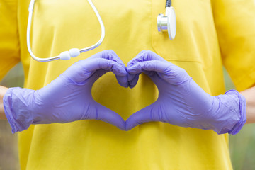 Detalle de manos de enfermera vestida con uniforme amarillo y guantes formando un corazón. - obrazy, fototapety, plakaty