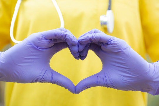 Detalle de manos de enfermera vestida con uniforme amarillo y guantes formando un coraz&oacute;n.