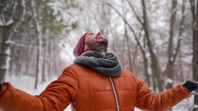 Hipster man enjoying falling snow outdoors on winter day on Christmas vacation. Happy guy open arms under snowfall in park and catch the snowflakes with hands and tongue. Male stretch out arms to sky.