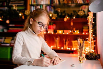 Adorable litle girl is writting a letter to Santa Klaus sitting on the table