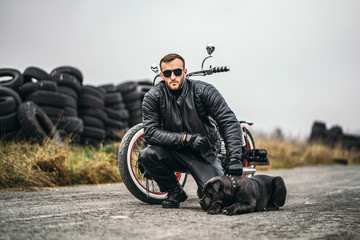 Obraz premium Biker in a leather suit crouched near his dog and red motorcycle on the road. Many tires on the background
