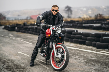 Bearded man in sunglasses and leather jacket looking at the camera while sitting on a motorcycle on the road. Behind him is a row of tires