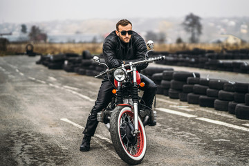 Naklejka premium Bearded man in sunglasses and leather jacket looking at the camera while sitting on a motorcycle on the road. Behind him is a row of tires