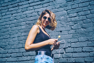 Portrait of positive hipster girl in sunglasses enjoying romantic playlist via earphones and media application on mobile phone, smiling woman with chupa chups posing near promotional background