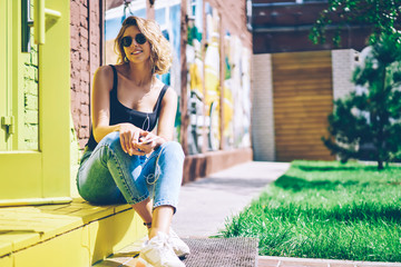 Portrait of happy hipster girl dressed in denim jeans sitting on urban setting and enjoying romantic music during sunny summer day, positive woman in sunglasses looking at camera while listening audio