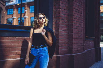 Charming caucasian model in sunglasses looking away and enjoying spring weather while walking on town streets with folder in hands, attractive female student in trendy apparel returning from college