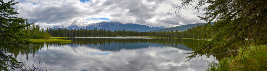 A view through the pine forest towards the snow capped mountains reflecting on the lake on a cloudy day.