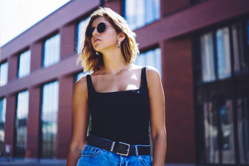 Serious attractive female student in stylish sunglasses looking away and thinking about free time during sunny summer day, beautiful female teenager casual dressed resting after walking in city