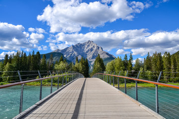 A wooden pedestrian bridge with wire tension cables and handrails on both sides with a view of a dense pine forest the rocky Cascade Mountain in the background on this partially cloudy day.