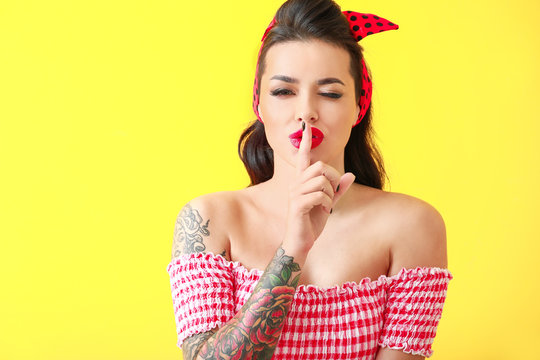 Portrait of beautiful tattooed pin-up woman showing silence gesture on color background