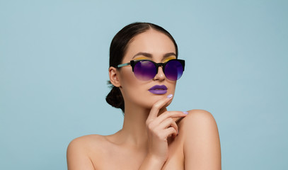 Fototapeta premium Portrait of beautiful woman with bright make-up and sunglasses on blue studio background. Stylish, fashionable make and hairstyle. Colors of summer. Beauty, fashion and ad concept. Serious, confident.