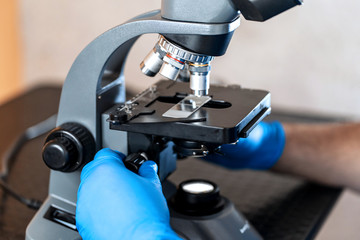 Male laboratory assistant examining biomaterial samples in a microscope. Cllose up hands in blue rubber gloves adjust microscope