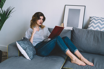 Interested young woman with eyeglasses in hand holding literature book and relaxing at home, pondering hipster girl reading bestseller lying on comfortable sofa in apartment with stylish interior