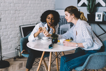 Surprised afro american female blogger feeling shocked from reading text message showed by caucasian friend sitting near, amazed women checking notification for smartphone gadget connected to 4g